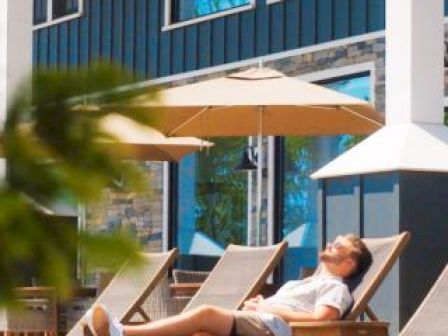 A person is relaxing on a lounge chair beside a pool with umbrellas and a modern building in the background.