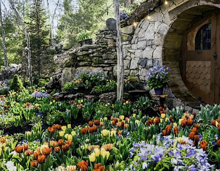 A circular wooden door set in a stone wall, surrounded by colorful tulips and greenery, reminiscent of a cozy, hobbit-like dwelling in a forest.