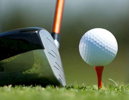 A close-up of a golf club poised to hit a golf ball on a tee on a golf course, with blurred background and green grass.