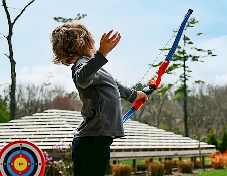 A child is using a toy bow and arrow at an outdoor archery range with colorful targets in a park on a clear day ending the sentence.