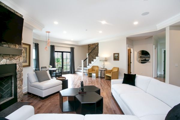 A spacious living room with white sofas, a fireplace, TV, coffee tables, dining area, stairs, and various decorations, featuring a modern interior design.