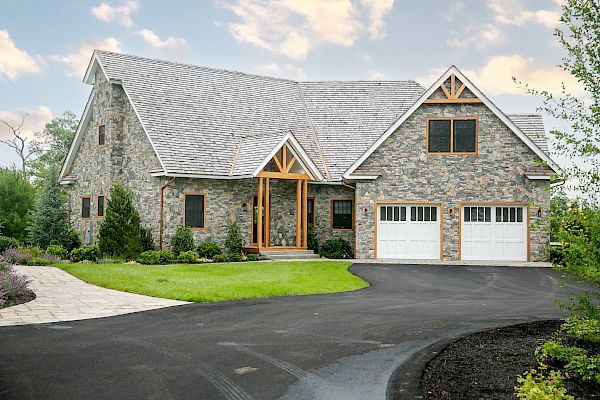 A stone house with a complex roof design, a well-maintained lawn, a double garage, and a paved driveway under a partly cloudy sky, ending the sentence.