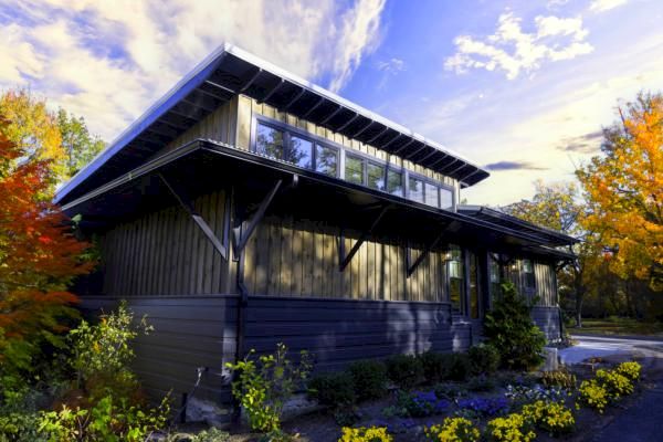 A modern house with large windows, surrounded by colorful autumn trees and a landscaped garden, under a partly cloudy sky.