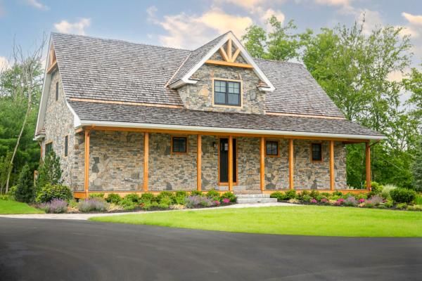 A stone house with a sloped roof, surrounded by greenery, featuring a front porch and wooden columns with a paved driveway leading to it.