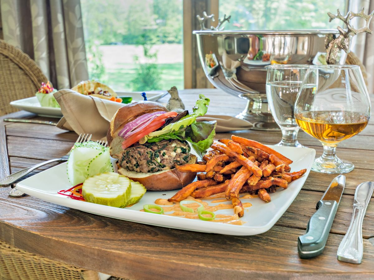 A burger with sweet potato fries, pickles, cucumber slices, and drinks on a wooden table with outdoor scenery in the background.