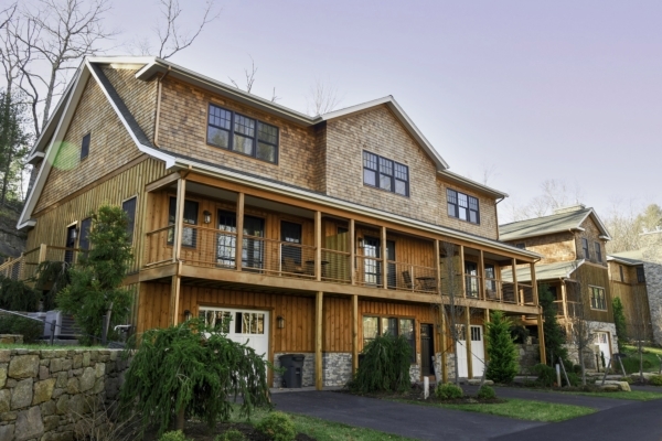 A large two-story house with a wooden exterior, featuring an expansive front porch, surrounded by greenery, and adjacent to a paved driveway.