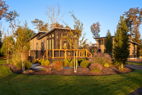 A modern cabin surrounded by lush landscaping and trees, with a wooden deck and large windows, under a clear blue sky.