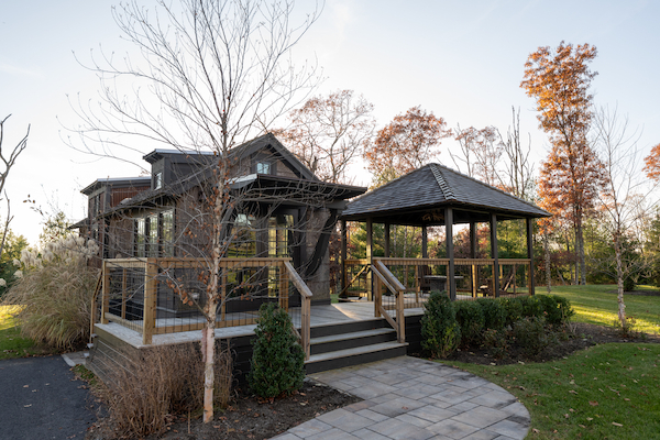 The image shows a modern rustic cabin with a porch and adjacent gazebo, surrounded by trees and a well-landscaped garden, on a sunny day.