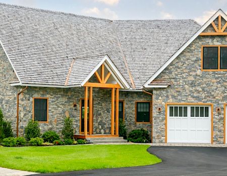 A beautiful stone-clad house with a large front yard, green landscaping, and a double garage with white doors, on a bright day with a partly cloudy sky.