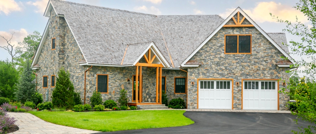 A beautiful stone-clad house with a large front yard, green landscaping, and a double garage with white doors, on a bright day with a partly cloudy sky.