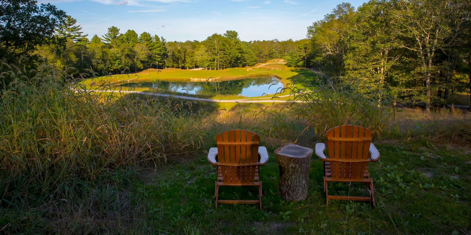 Two wooden chairs and a small table overlook a scenic pond surrounded by trees and greenery under a clear blue sky, ending the sentence.