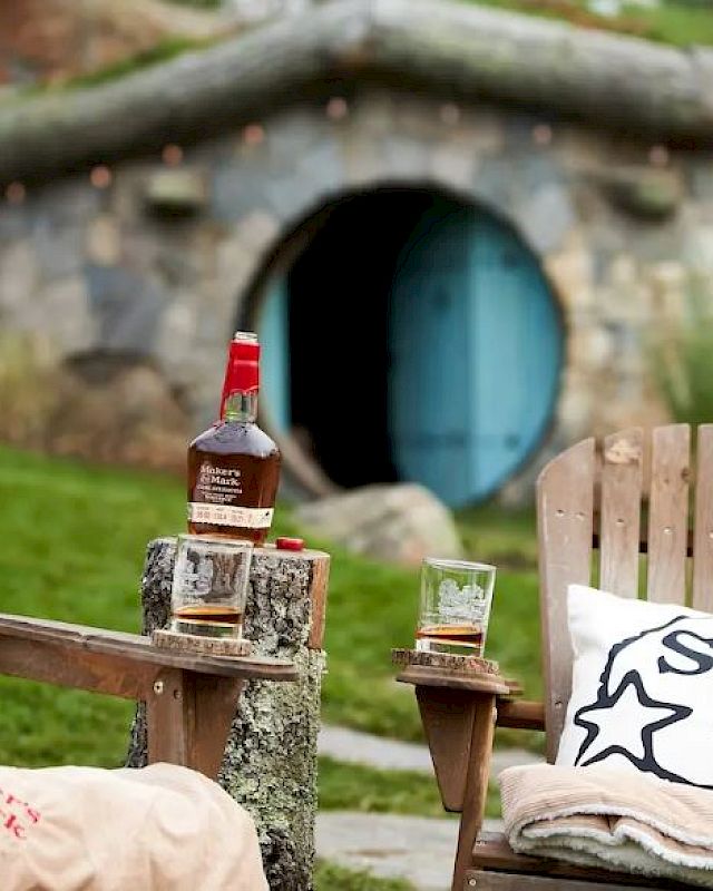 Two wooden chairs with pillows and blankets, a table holding a bottle of liquor and two glasses, in front of a Hobbit-style house entrance.