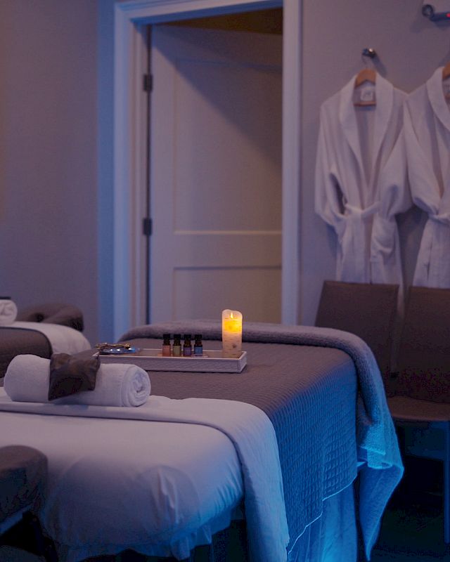 A spa room with two massage tables, a lit candle, rolled towels, two bathrobes on a hook, a sink, and soft lighting creating a relaxing ambiance.