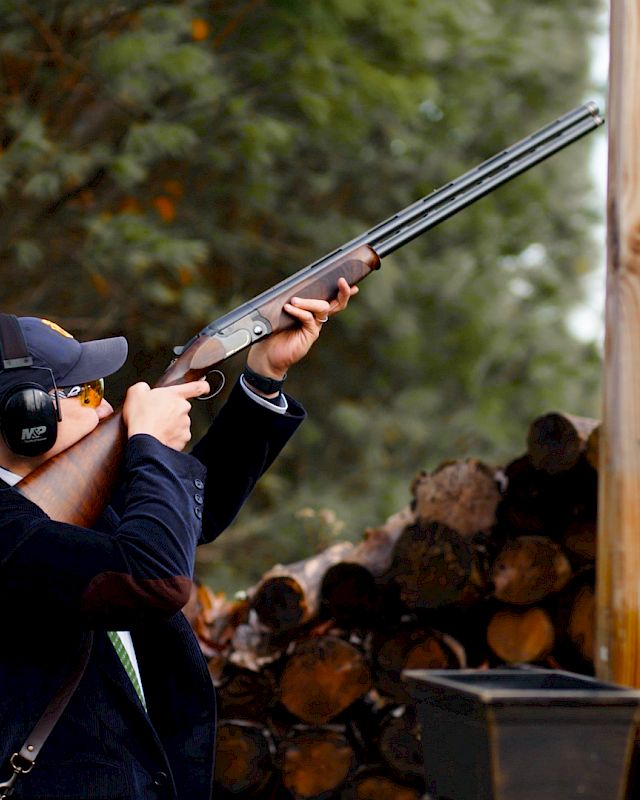 A person wearing protective earmuffs and shooting eyewear is aiming a shotgun at an outdoor shooting range, surrounded by logs and trees.