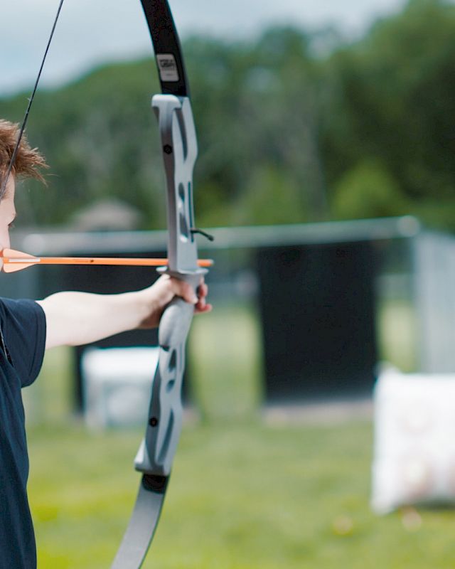 A person is aiming a bow and arrow at a target in an outdoor archery range, with greenery and other targets in the background.