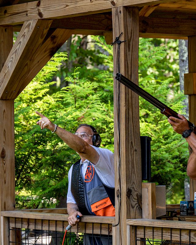 Two men participating in clay pigeon shooting; one is aiming a shotgun while the other is pointing in a direction, under a wooden shelter structure.