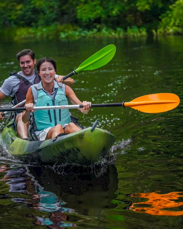 Two people are kayaking on a calm body of water, smiling and paddling. The kayak has vibrant green and orange paddles.