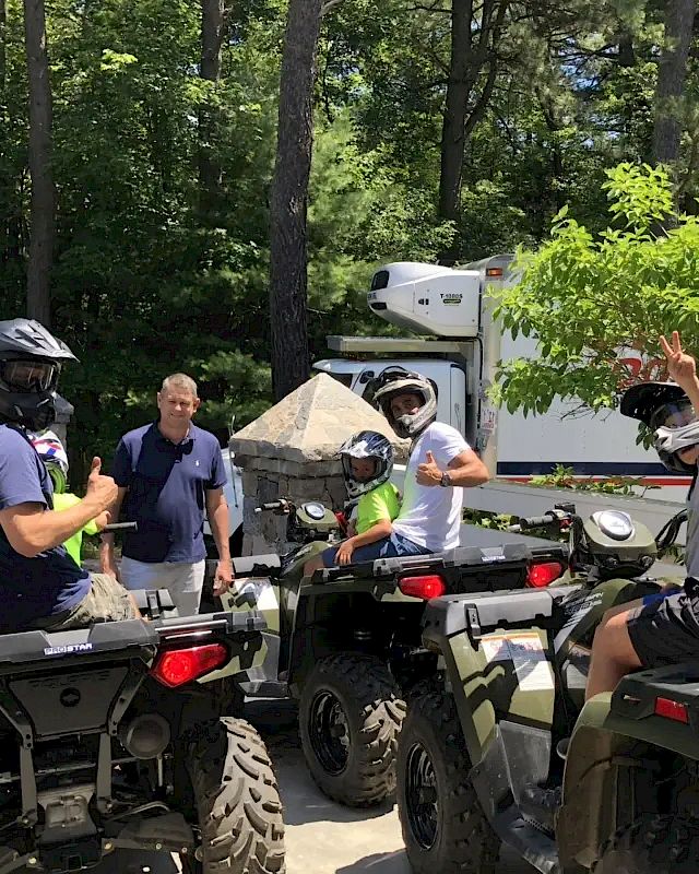 A group of people wearing helmets and sitting on ATVs while a man stands nearby in a wooded area.