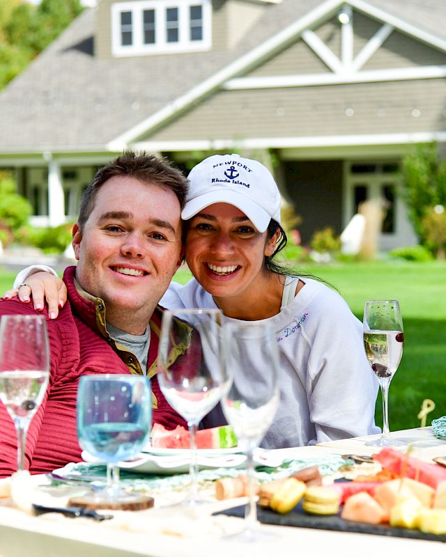 A smiling couple enjoys an outdoor meal with various dishes and drinks on a table, in front of a house with a yard.