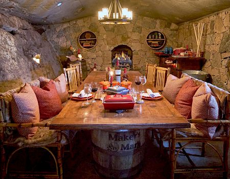 A cozy, rustic dining room with stone walls, wooden furniture, and a central table set for dining, featuring cushioned chairs and warm lighting.