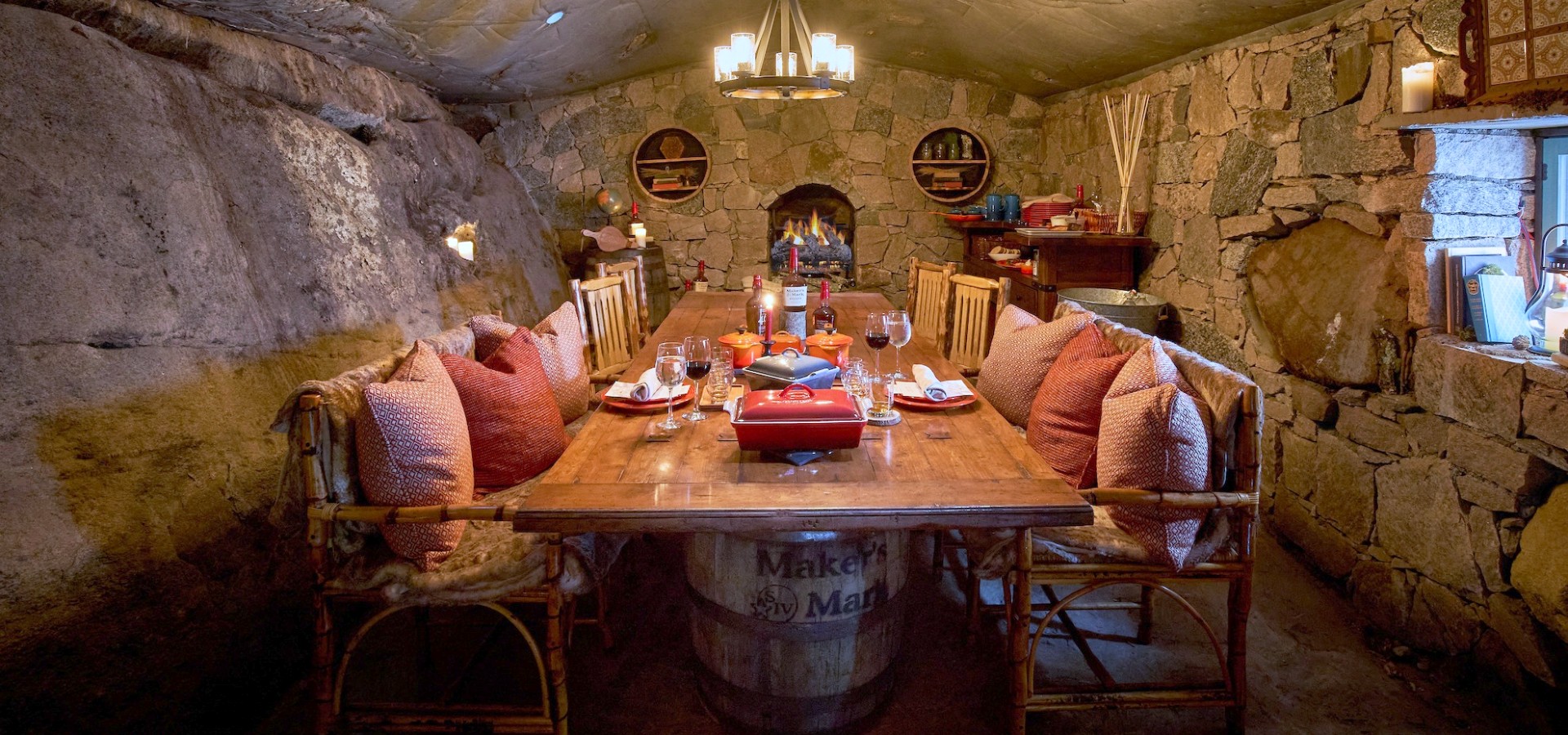 A cozy, rustic dining room with stone walls, wooden furniture, and a central table set for dining, featuring cushioned chairs and warm lighting.