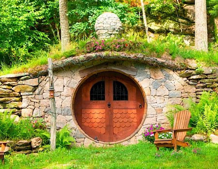 A charming, hobbit-like house with a round wooden door set in a grassy hillside, flanked by two wooden chairs, and lush green surroundings.