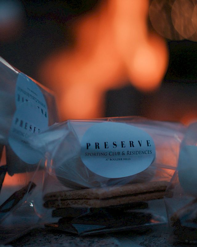 Packaged s'mores kits with graham crackers, marshmallows, and chocolate are in focus, with a blurred fire in the background.