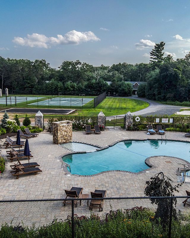 The image shows an outdoor recreational area with a swimming pool, sun loungers, tennis courts, and lush greenery surrounding the space.