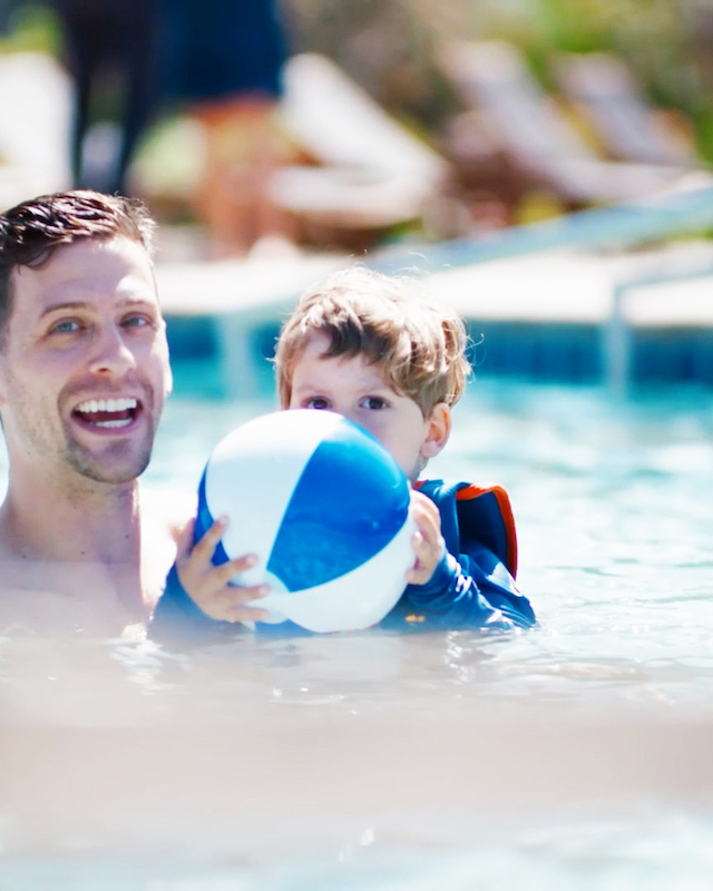 A man and a child are playing in a swimming pool, with the child holding a blue and white beach ball. The background shows blurred pool chairs.