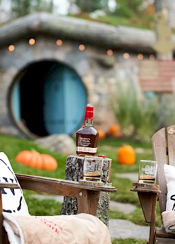 A cozy outdoor setting with wooden chairs, blankets, a bottle of liquor, glasses, pumpkins, and a round hobbit-like house entrance in the background.