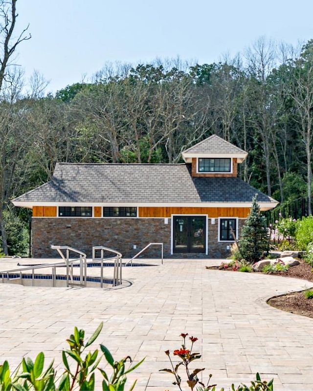 The image shows a small stone and wood building with a gabled roof, surrounded by trees and pathways. There are metal handrails near the entrance.