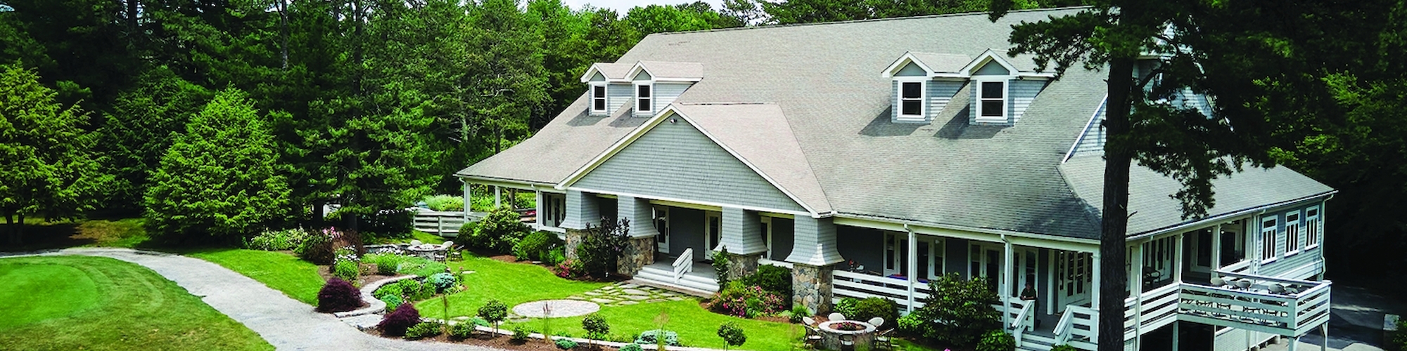 This image shows a large house with a gray roof surrounded by well-maintained lawns, trees, and paths under a partly cloudy sky.