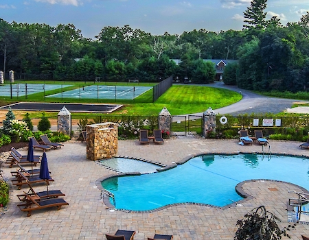 This image features an outdoor swimming pool area with lounge chairs and umbrellas, adjacent to tennis courts, surrounded by lush greenery and trees.