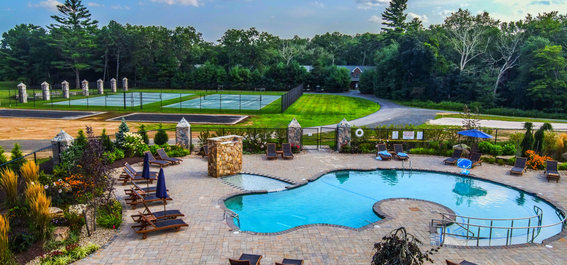 This image features an outdoor swimming pool area with lounge chairs and umbrellas, adjacent to tennis courts, surrounded by lush greenery and trees.