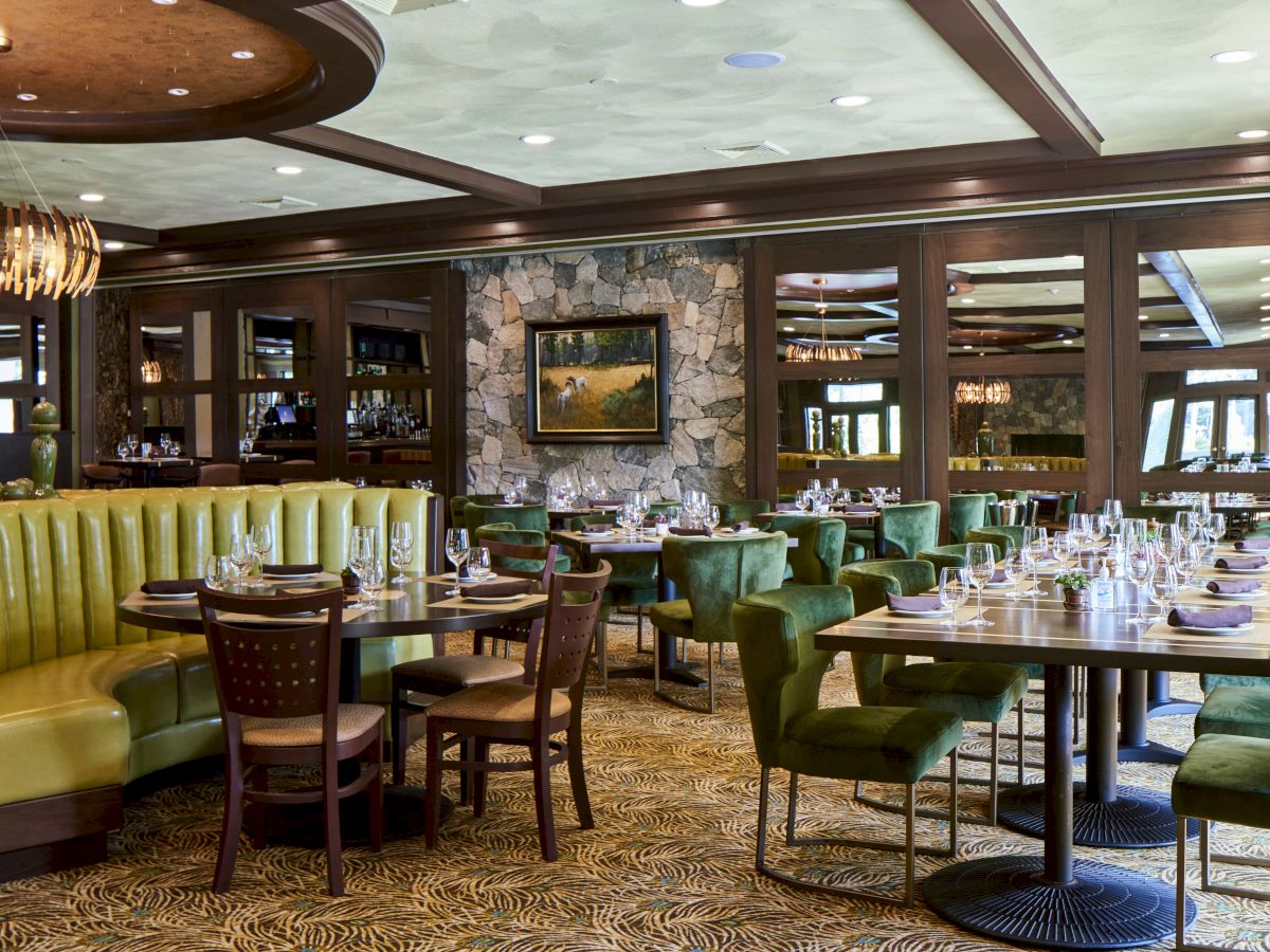 A cozy restaurant interior with green cushioned chairs, wooden tables set with glassware, a stone-paneled wall, and artistic decor.