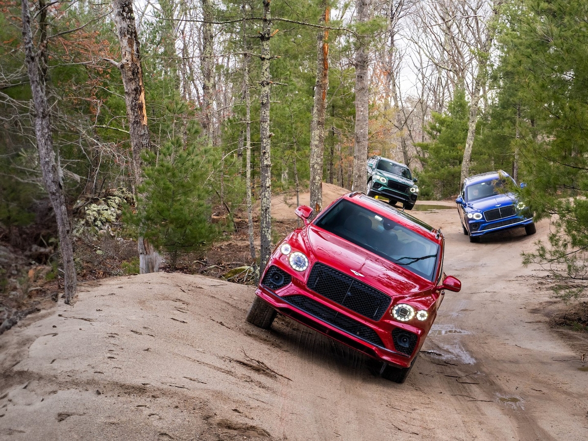 Three SUVs navigate a dirt trail in a forested area, with a red one leading, followed by green and blue ones in the background.