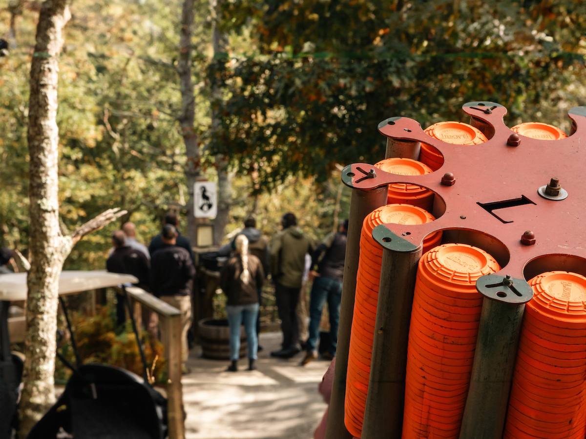 The image shows a clay pigeon shooting range with a clay target thrower in focus and people gathered in the background in a wooded area.