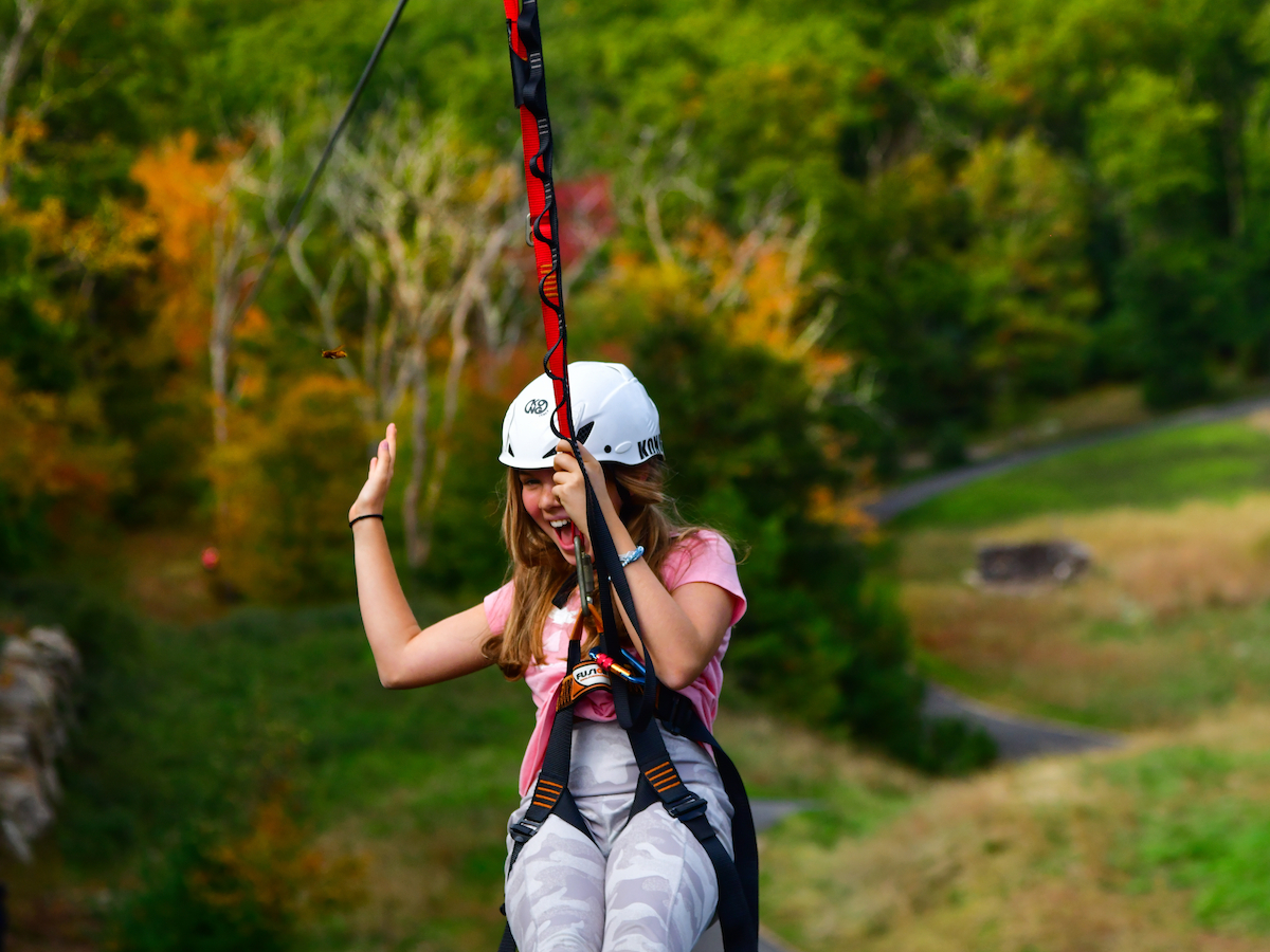 A person wearing a helmet is zip-lining over a lush, green landscape with a winding path below and trees displaying autumn colors.