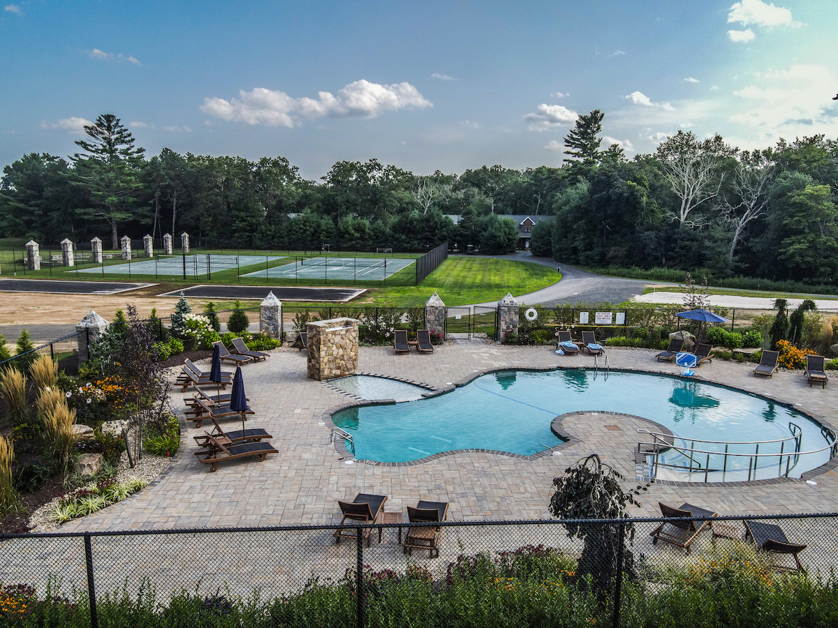 This image shows an outdoor area featuring a swimming pool, lounge chairs, tennis courts, and landscaped surroundings with trees in the background.