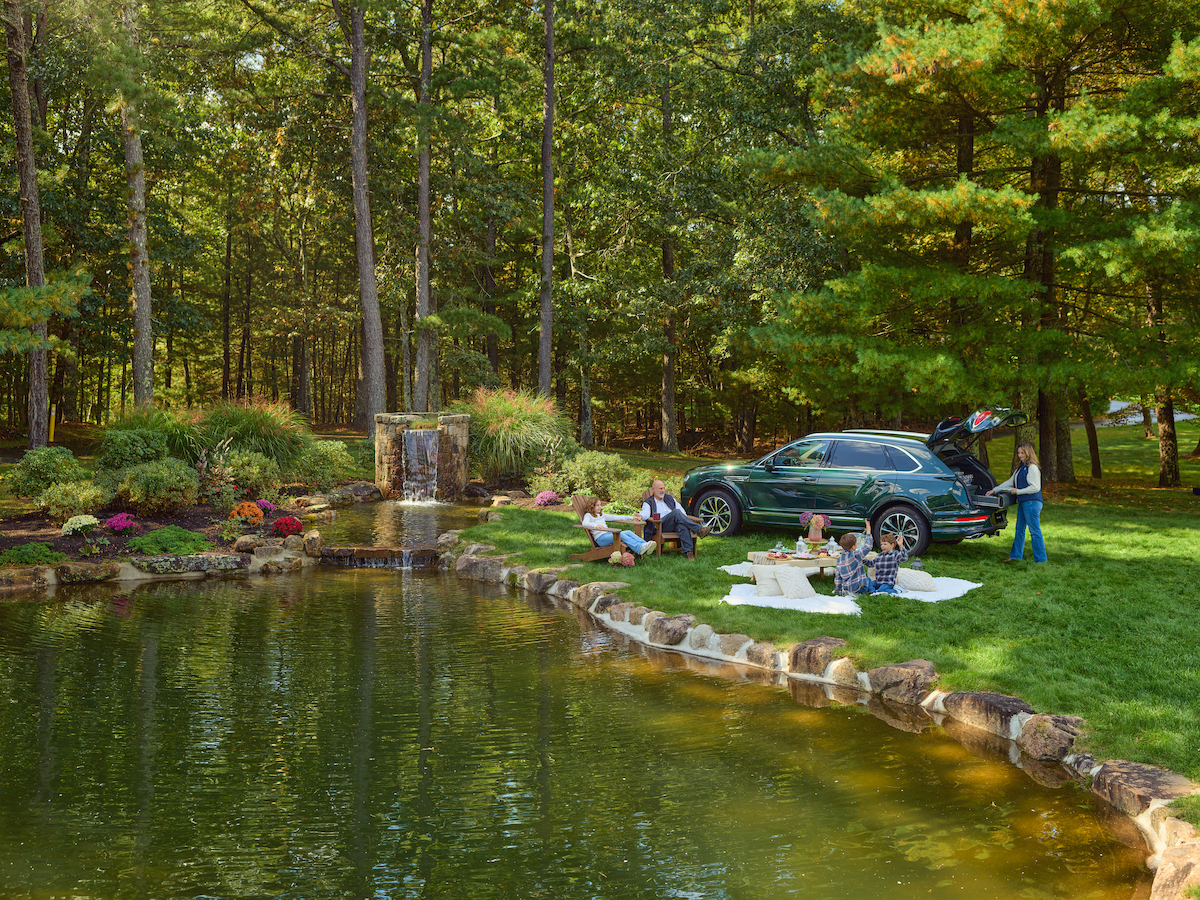 A group of people is having a picnic by a pond in a wooded area with a waterfall, next to a parked SUV with its trunk open.