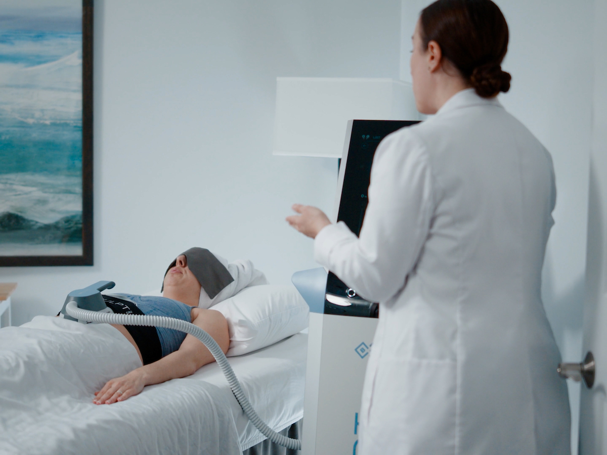 A patient is lying on a medical bed with a device attached to their abdomen, attended by a healthcare professional in a white coat, in a clinical setting.