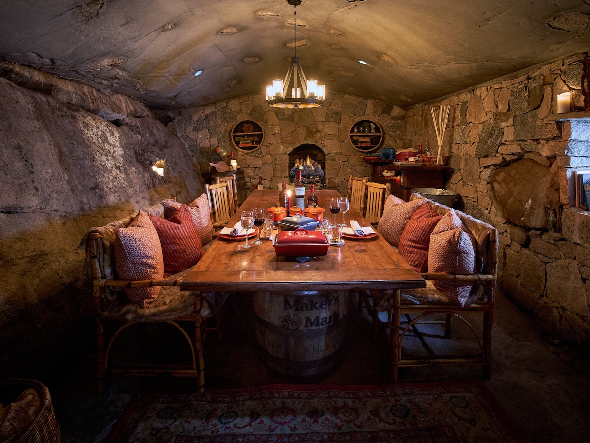 A cozy stone-walled dining room with a wooden table, cushioned chairs, warm lighting, and various decorative items is shown in the image.