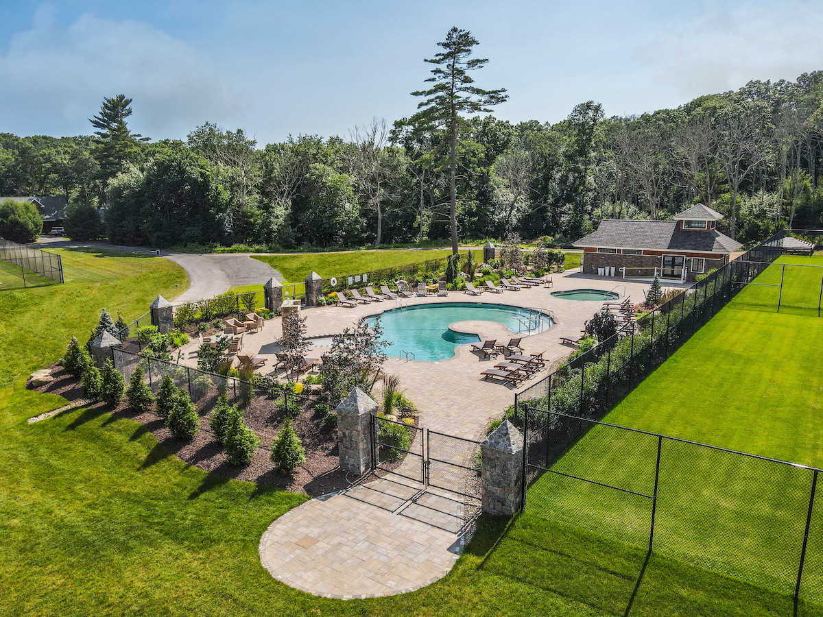 The image shows an outdoor pool with surrounding lounge chairs and landscaping, enclosed by a fence, with a building nearby and greenery in the background.