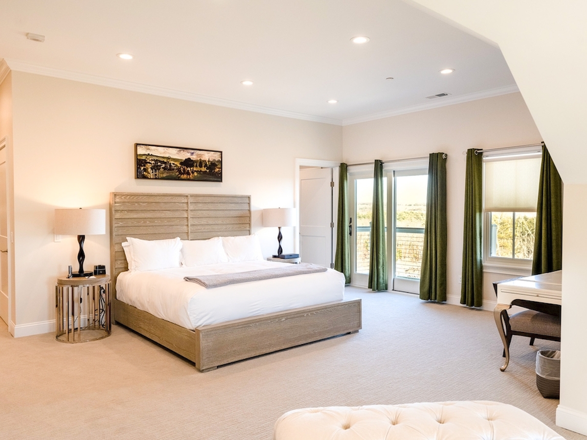 A spacious bedroom with a large bed, bedside tables, lamps, green curtains, a desk by the windows, and a white ottoman in the foreground.