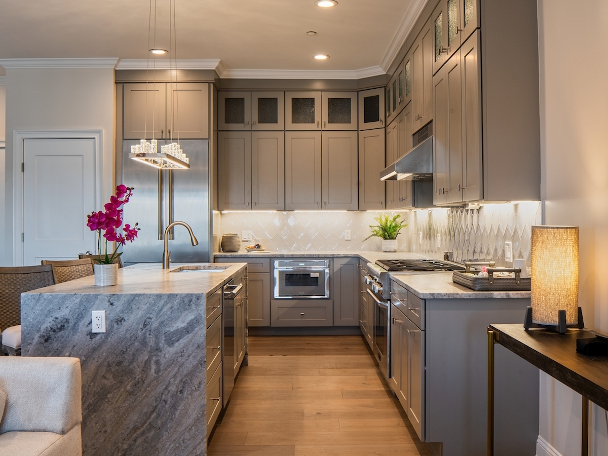 A modern kitchen with gray cabinets, a marble island, and stainless steel appliances. There are decorative plants and a lamp on a side table.