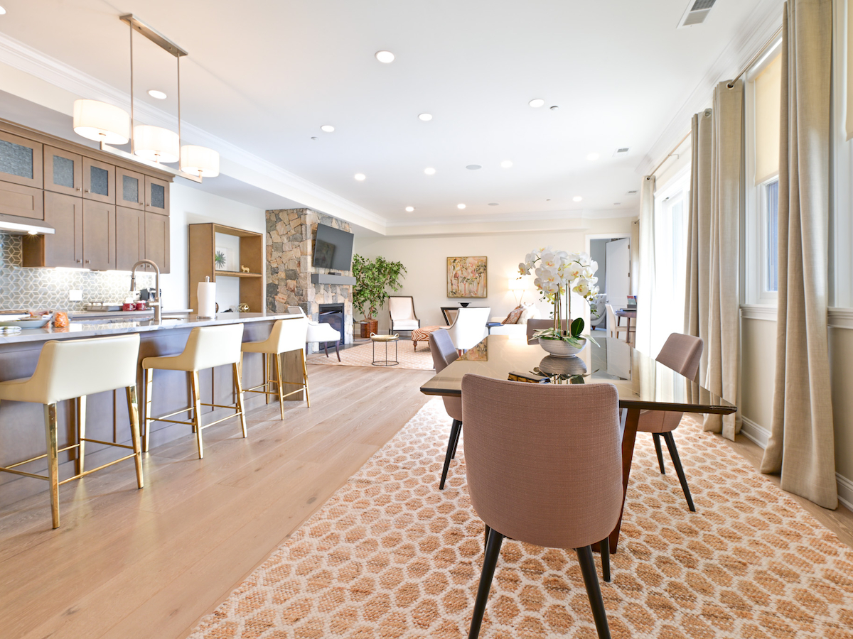 An open-concept kitchen and living area featuring modern decor, a dining table, high chairs at the kitchen island, and large windows allowing natural light.