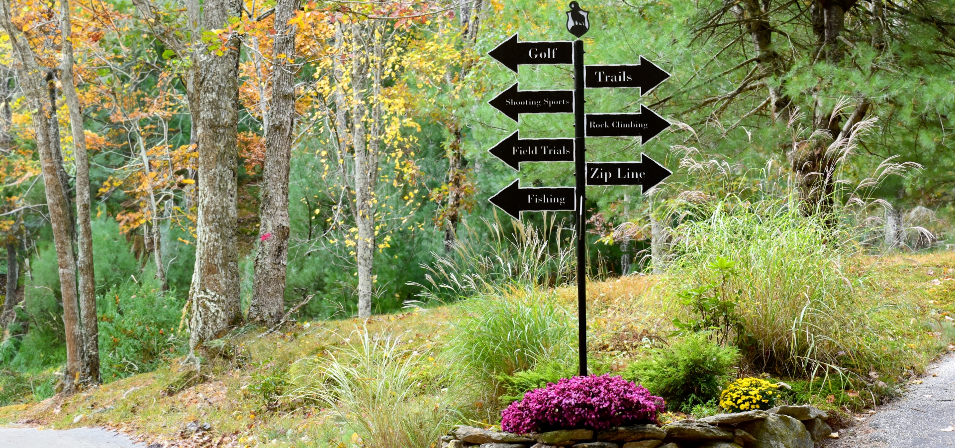 A signpost in a forested area has multiple direction signs for Golf, Trails, Mountain Sports, Fish Trails, Black & Blue Lake, Zip Line, and Fishing.