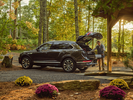 A person unloads items from the trunk of a black SUV parked in a forest setting near a wooden structure decorated with colorful flowers.