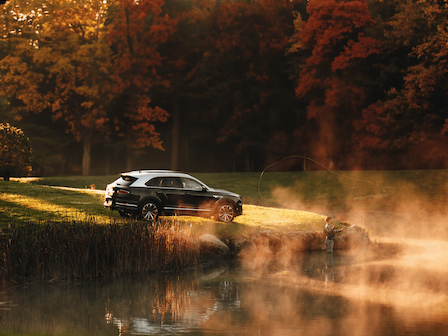 A person fishing by a misty pond next to a parked car in a scenic autumn forest with colorful foliage.