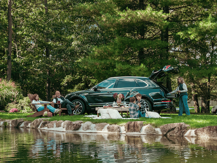 A group of people having a picnic by a pond with a parked SUV and a forested area in the background, enjoying the outdoors.